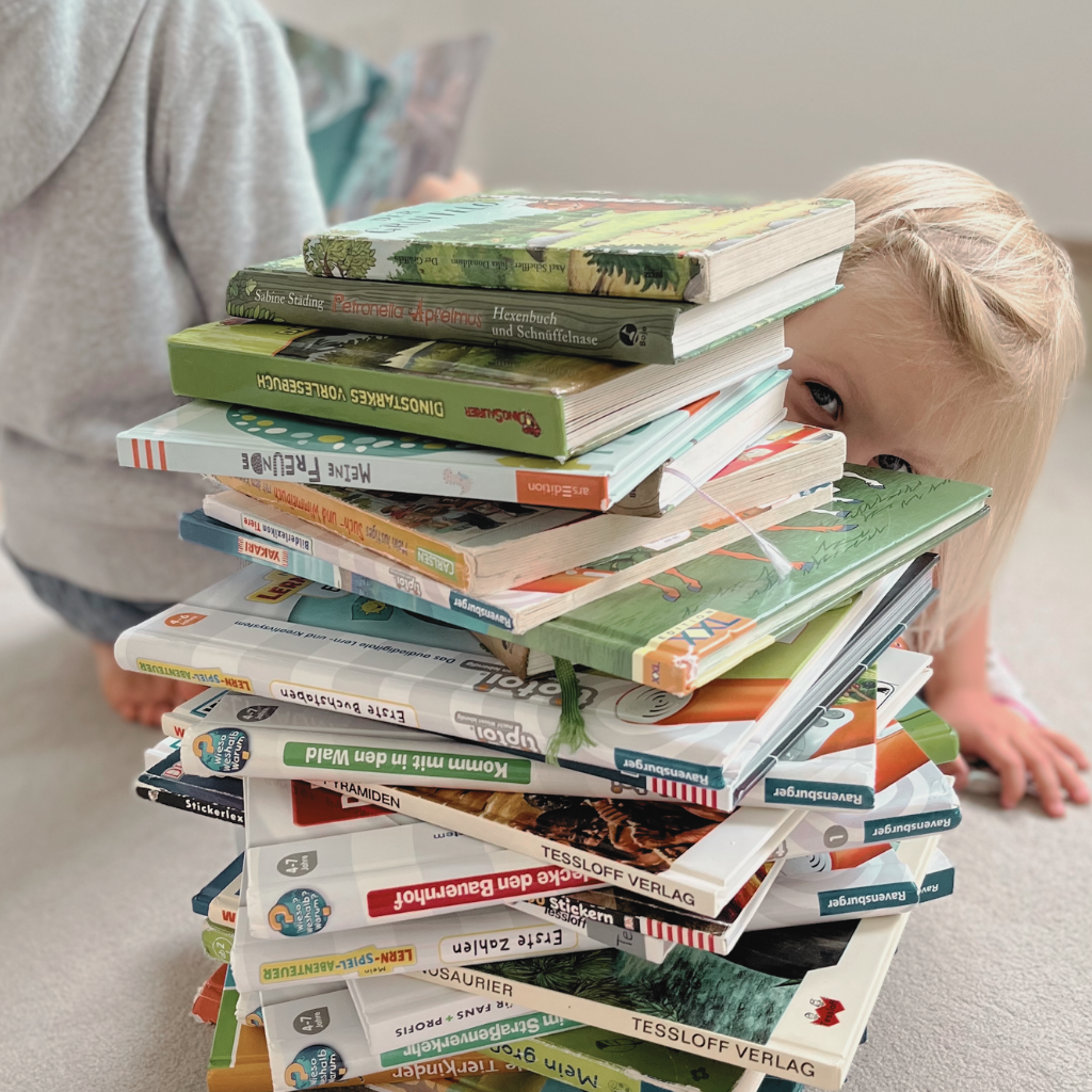 Kleines Mädchen schaut hinter einem hohen Stapel verschiedener Kinderbücher hervor